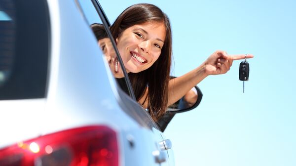 Junge Frau lehnt sich aus dem Autofenster und hält lachend einen Autoschlüssel in der Hand.