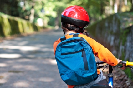 Ein Kind fährt auf einem Fahrrad eine Straße entlang. Man sieht es von hinten. Es trägt einen roten Fahrradhelm und eine blaue Schultasche am Rücken.