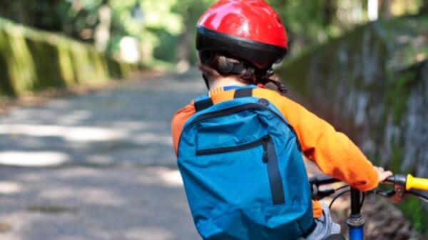 Ein Kind fährt auf einem Fahrrad eine Straße entlang. Man sieht es von hinten. Es trägt einen roten Fahrradhelm und eine blaue Schultasche am Rücken.