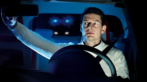 Ein junger Lenker im Auto schaut in den Rückspiegel. Hinter der Heckscheibe sieht man ein Fahrzeug mit Blaulicht.