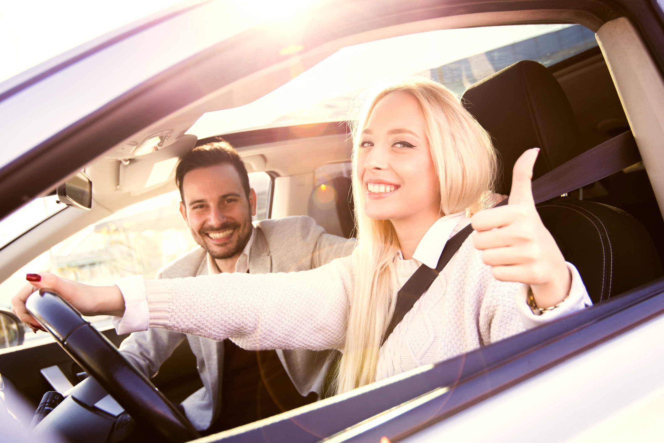 Ein junges, lachendes Pärchen befindet sich im Auto; die junge Frau hält eine Hand am Lenkrad, mit der anderen gibt sie ein "Daumen hoch"-Zeichen.