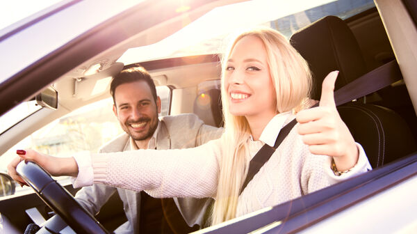 Ein junges, lachendes Pärchen befindet sich im Auto; die junge Frau hält eine Hand am Lenkrad, mit der anderen gibt sie ein "Daumen hoch"-Zeichen.