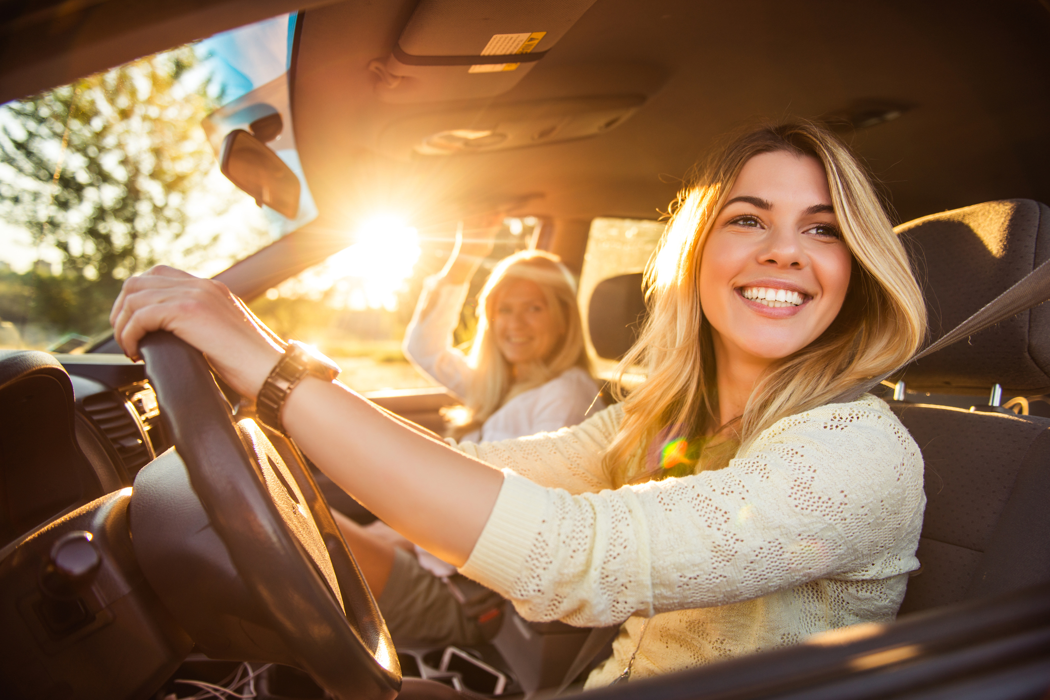 Junge Frau am Steuer im Auto bei sonnigem Wetter unterwegs. Sie schaut nach links und lacht. Am Beifahrersitz befindet sich eine weitere Frau.