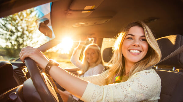 Junge Frau am Steuer im Auto bei sonnigem Wetter unterwegs. Sie schaut nach links und lacht. Am Beifahrersitz befindet sich eine weitere Frau.