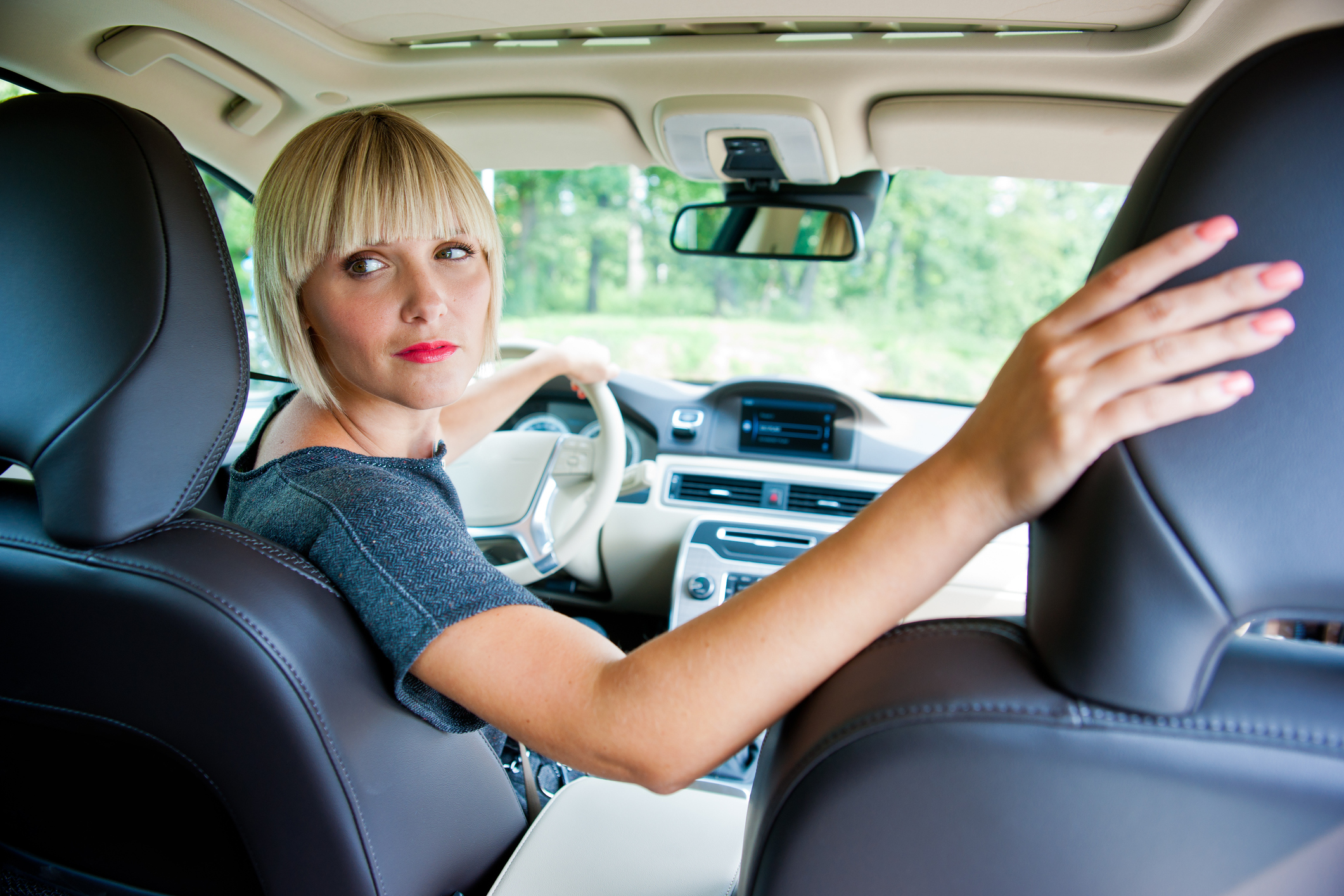 Eine Frau mit blonden Haaren sitzt im Auto am Steuer und schaut über die rechte Schulter nach hinten. Sie trägt ein graues T-Shirt. Der rechte Arm liegt auf der Beifahrersitzlehne.