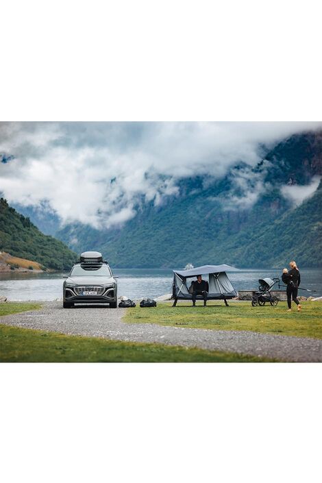 Ein Auto parkt auf einem Weg vor einem See in den Bergen. EIn paar Meter entfernt ist das Thule Outset aufgebaut. Der Regenschutz ist angebracht und ein Mann sitzt seitlich bei einer Öffnung und eine Frau geht mit einem Kind in ihrem Arm auf das Zelt zu.