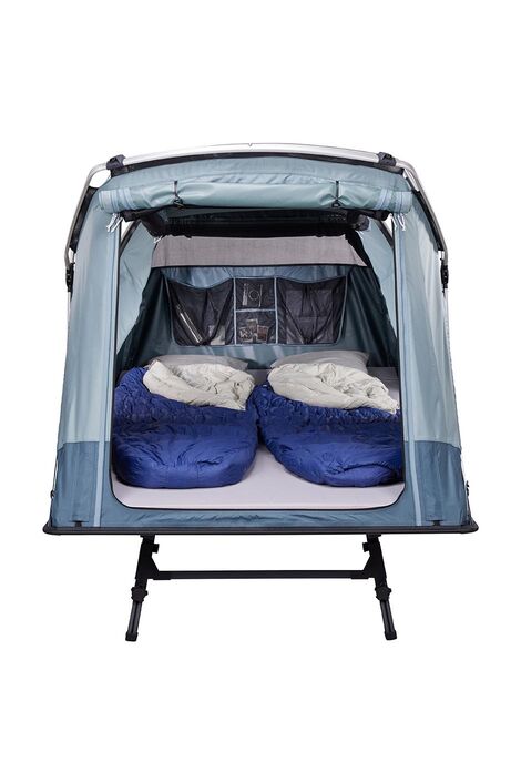 Frontal Sicht auf das Zelt. Der Eingang ist offen und im inneren liegen 2 Schlafsäcke. An der hinteren Wand sieht man mehrere Taschen in denen ein paar Gegenstände verstaut sind.