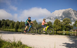 Zwei Radfahrer fahren auf einem Radweg an einem Fluss, im Hintergrund ein Berg