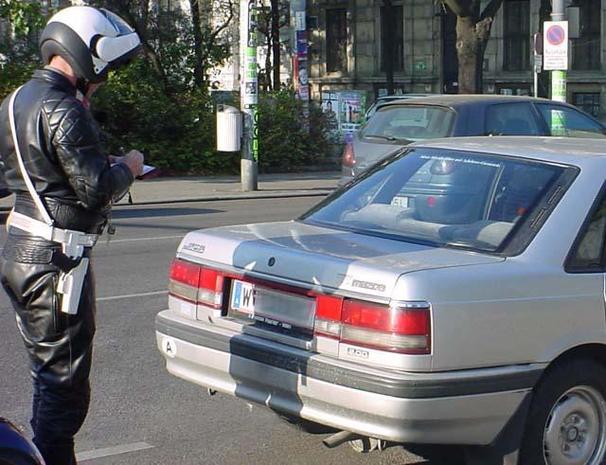 Ein Polizist in schwarzer Motorradbekleidung mit Helm am Kopf steht hinter einem silbernen Auto und stellt einen Strafzettel aus.
