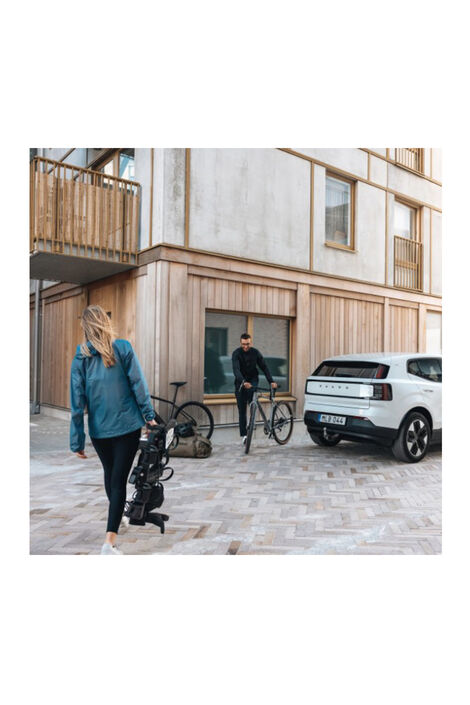 16989 Outpace 2er 5 g Eine Frau trägt den Fahrradträger zu einem weißen Auto. Ein Mann schiebt ein Fahrrad zum Auto und ein Fahrrad lehnt neben ihnen an der Hauswand.
