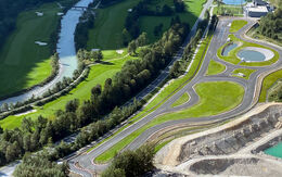 Inmitten einer ländlichen Landschaft erstreckt sich das Experience Center Saalfelden/Brandlhof. Eine große asphaltierte Übungsfläche, mit Kreuzungselementen und Übungsstraßen. Links der Übungsfläche sind eine Bundesstraße, Wiesen, ein Fluss mit zwei Brücken und ein anschließender Wald.