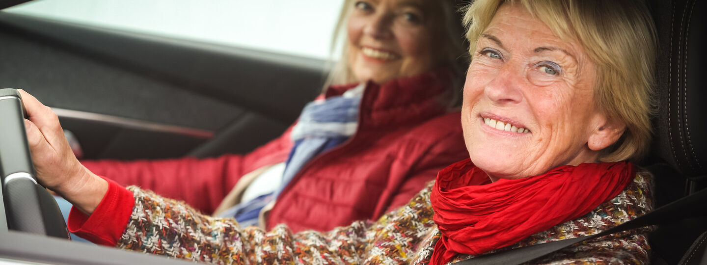 Zwei lächelnde Frauen sitzen in einem roten Auto. Die Fahrerin trägt einen bunten Pullover und einen roten Schal, die Beifahrerin eine rote Jacke und einen blauen Schal.