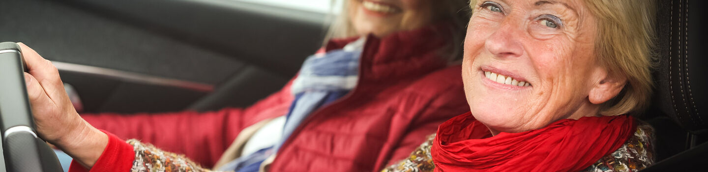 Zwei lächelnde Frauen sitzen in einem roten Auto. Die Fahrerin trägt einen bunten Pullover und einen roten Schal, die Beifahrerin eine rote Jacke und einen blauen Schal.