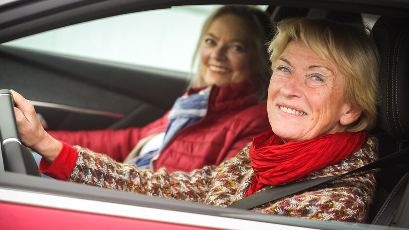 Zwei lächelnde Frauen sitzen in einem roten Auto. Die Fahrerin trägt einen bunten Pullover und einen roten Schal, die Beifahrerin eine rote Jacke und einen blauen Schal.