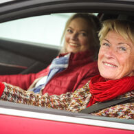 Zwei lächelnde Frauen sitzen in einem roten Auto. Die Fahrerin trägt einen bunten Pullover und einen roten Schal, die Beifahrerin eine rote Jacke und einen blauen Schal.