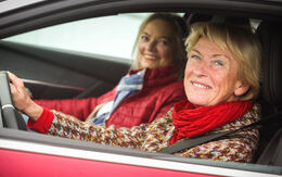 Zwei lächelnde Frauen sitzen in einem roten Auto. Die Fahrerin trägt einen bunten Pullover und einen roten Schal, die Beifahrerin eine rote Jacke und einen blauen Schal.