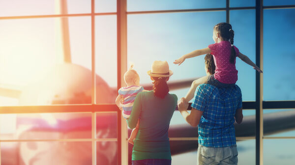 Eine Familie mit zwei Kindern schaut durch ein Flughafenfenster auf ein Flugzeug.