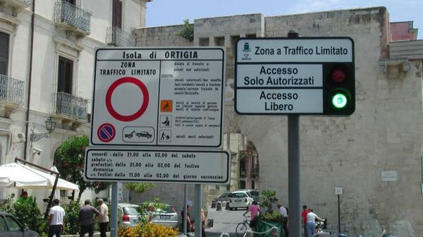 Zwei Schilder in einer italienischen Stadt. Das linke Schild weist auf eine Zone mit beschränktem Verkehr (Zona Traffico Limitato) in Ortigia hin, mit einem Durchfahrtsverbotsschild. Das rechte Schild hat eine Ampel und die Aufschriften "Accesso Solo Autorizzati" und "Accesso Libero". Im Hintergrund sind Gebäude, Autos und Menschen zu sehen.