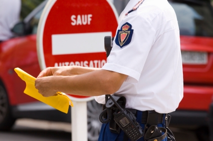 Ein Polizist, der ein gelbes Dokument oder Formular in der Hand hält. Im Hintergurnd ist ein rotes Verbotsschild mit einem weißen Balken und der Aufschrift "SAUF AUTORISATION" zu sehen. Ein rotes Auto ist ebenfalls im Hintergrund zu erkennen.