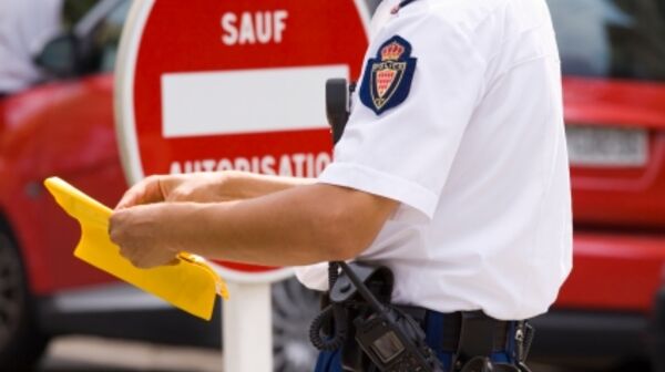Ein Polizist, der ein gelbes Dokument oder Formular in der Hand hält. Im Hintergurnd ist ein rotes Verbotsschild mit einem weißen Balken und der Aufschrift "SAUF AUTORISATION" zu sehen. Ein rotes Auto ist ebenfalls im Hintergrund zu erkennen.