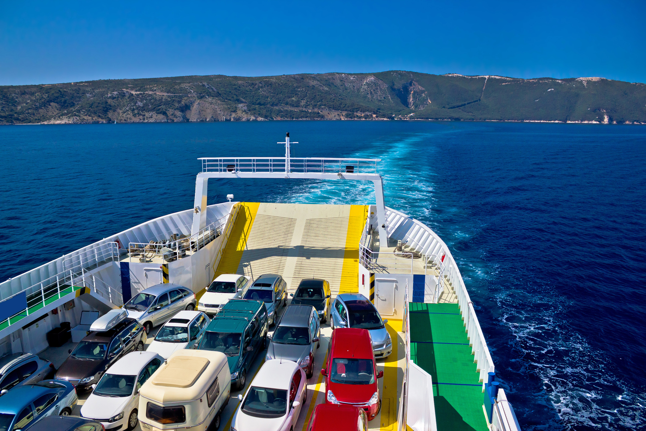 Eine Autofähre fährt gerade mit vielen unterschieldichen Autos beladen im Meer. Im Hintergrund sieht man hügeliges Land.