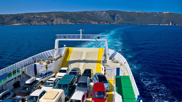 Eine Autofähre fährt gerade mit vielen unterschieldichen Autos beladen im Meer. Im Hintergrund sieht man hügeliges Land.