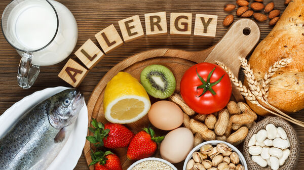 Auf einem Holztisch befinden sich ein Fisch, eine Kanne Milch, Erdbeeren, Nüsse, ein Brot, Eier, eine Kiwi, eine Tomate, Bohnen, eine Zitrone und Sesam. Über dem Brett bilden Holzbuchstaben das Wort "ALLERGY".
