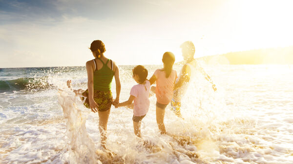 Eine Jugendliche und drei Kinder halten sich an den Händen an einem Strand und plantschen mit ihren Beinem im Meer.