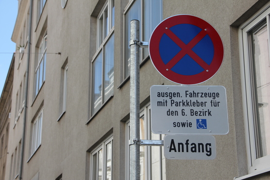 Das Bild zeigt ein Parkverbotsschild mit zwei Zusatztafeln in Wien. Auf dem oberen Zusatzschild steht: "ausgen. Fahrzeuge mit Parkkleber für den 6. Bezirk sowie" und zeigt ein Symbol für einen Behindertenparkplatz. Das untere Zusatzschild zeigt das Wort "Anfang“.
