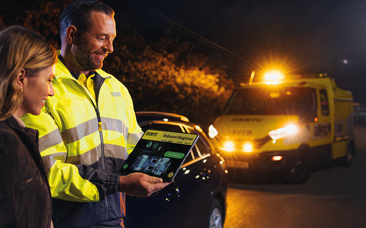 Ein ÖAMTC Pannenhelfer mit gelber Warnjacke hält ein Tablet in der Hand und zeigt es der Frau eben ihm. Es ist dunkel, die zwei Menschen stehen am Straßenrand und im Hintergrund steht ein Auto sowie ein Pannenhilfefahrzeug mit leuchtenden Scheinwerfern.