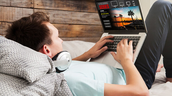 Ein junger Mann liegt im Bett, trägt Kopfhörer um den Hals und schaut auf einem Laptop eine Streaming-Serie.