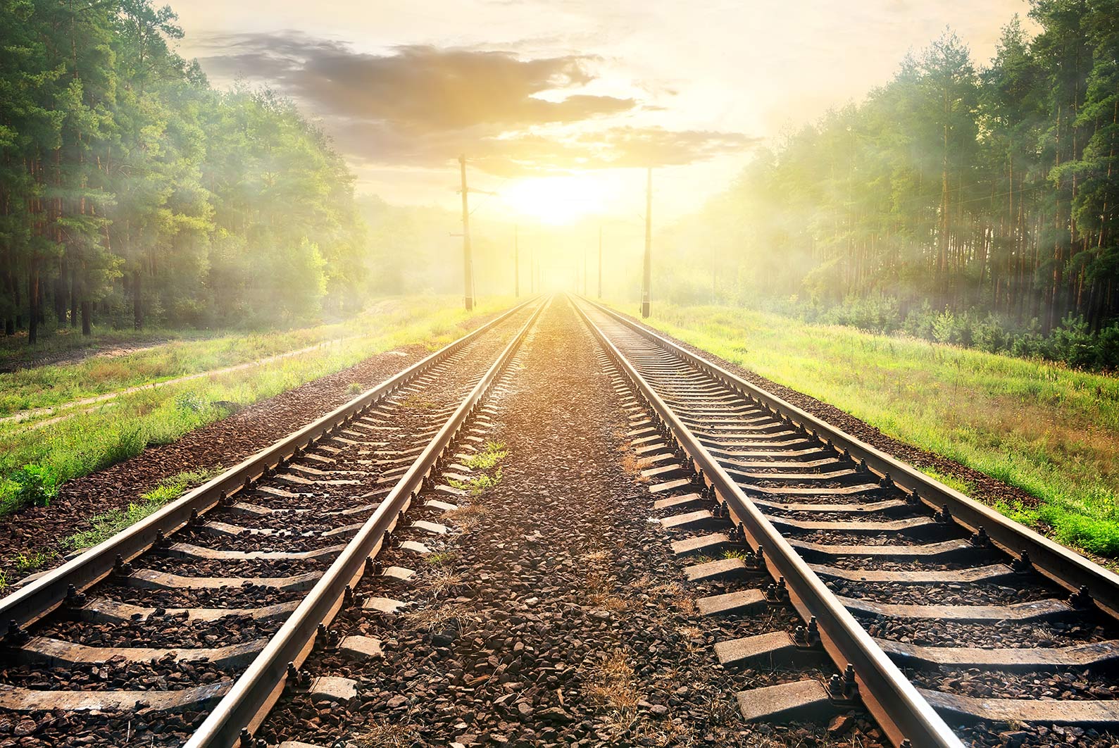 Zwei Eisenbahnschienen verlaufen parallel durch eine grüne Landschaft in Richtung eines Sonnenaufgangs oder Sonnenuntergangs am Horizont.