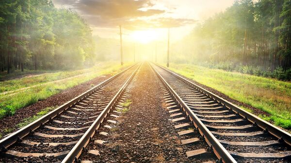 Zwei Eisenbahnschienen verlaufen parallel durch eine grüne Landschaft in Richtung eines Sonnenaufgangs oder Sonnenuntergangs am Horizont.