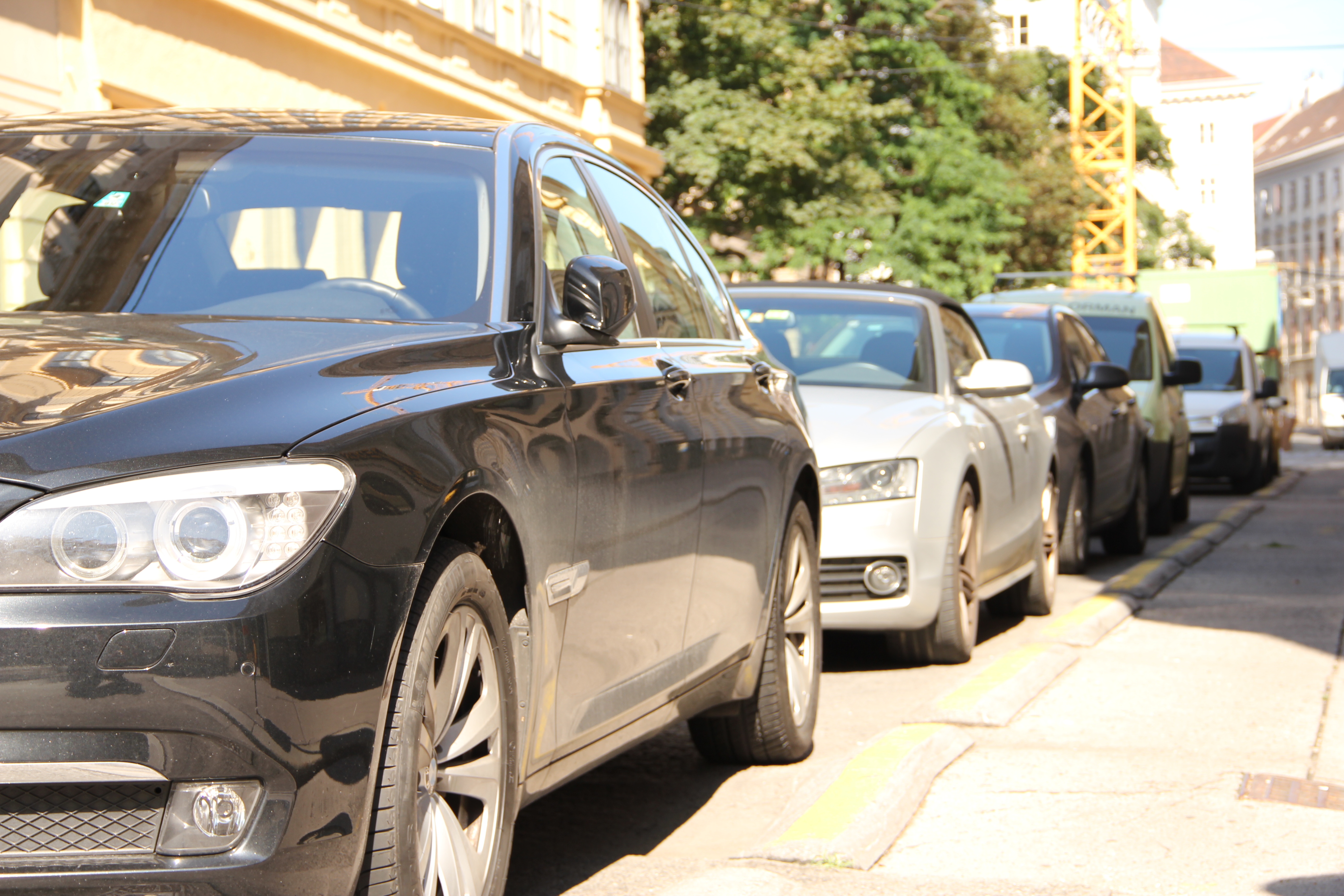 Parkende Autos stehen am Straßenrand einer Kurzparkzone im Sonnenschein. Im Hintergrund sind Mietshäuser, ein großer Baum und ein Kran sichtbar.