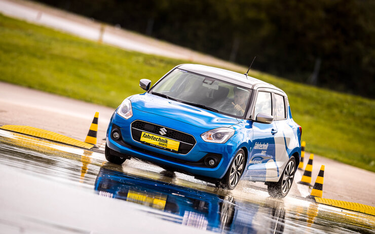 Ein blaues Auto fährt auf nassem Asphalt. Im Hintergrund sind gelb-schwarze Hütchen und grüne Wiesen.