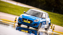 Ein blaues Auto fährt auf nassem Asphalt. Im Hintergrund sind gelb-schwarze Hütchen und grüne Wiesen.