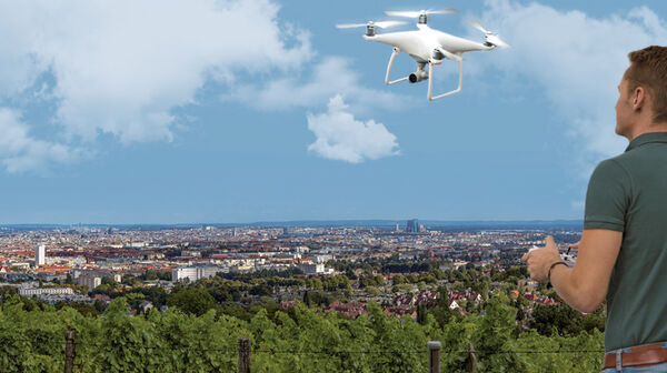 Ein Mann steuert eine weiße Drohne, die vor ihm in der Luft fliegt. Er steht auf einem Aussichtplatz am Rande einer Großstadt.