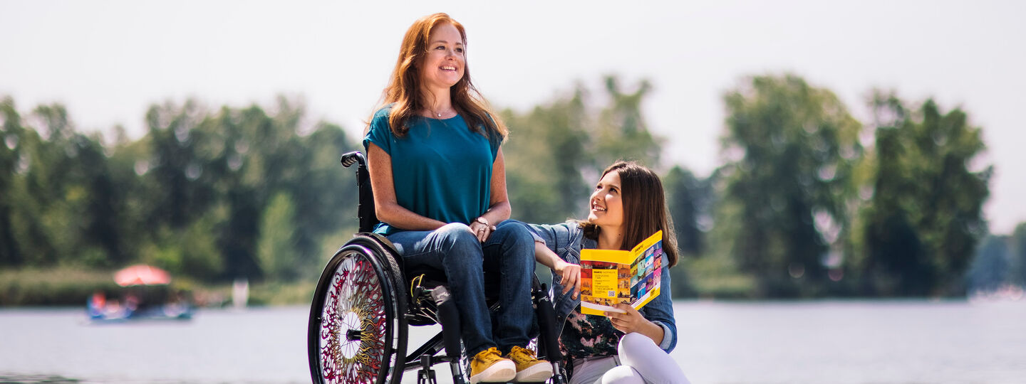 Eine Frau mit langen rotbraunen Haaren sitzt in einem Rollstuhl. Neben ihr sitzt eine Frau am Boden und hält ein Prospekt. Im Hintergrund sieht man unscharf Bäume und Wasser.