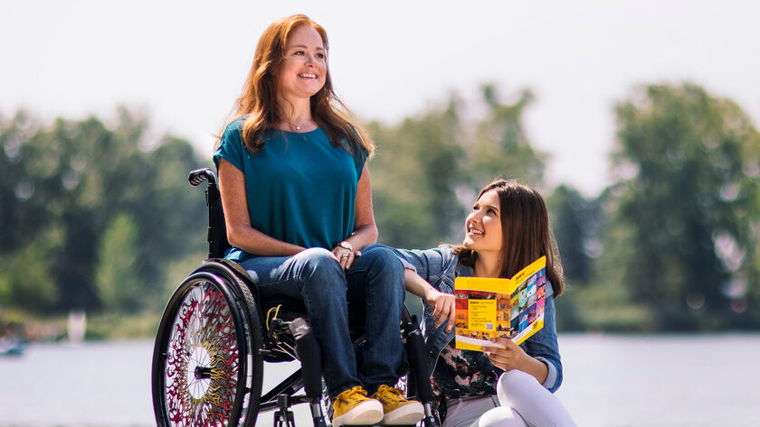 Eine Frau mit langen rotbraunen Haaren sitzt in einem Rollstuhl. Neben ihr sitzt eine Frau am Boden und hält ein Prospekt. Im Hintergrund sieht man unscharf Bäume und Wasser.