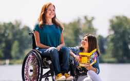 Eine Frau mit langen rotbraunen Haaren, blauer Kleidung und gelben Schuhen sitzt in einem Rollstuhl. Neben ihr sitzt eine Frau in Jeansjacke und weißer Hose am Boden und hält ein Prospekt. Im Hintergrund sieht man unscharf Bäume und Wasser.