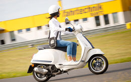 Eine Frau mit Helm fährt auf einer weißen Vespa. Im Hintergrund ist ein gelb weißes Gebäude mit Fahrtechnik Aufschrift.
