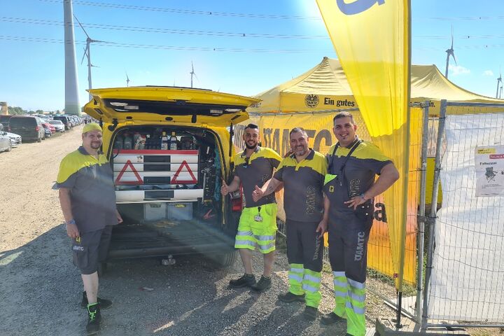 Vier Männer in ÖAMTC-Uniformen stehen vor einem gelben ÖAMTC-Fahrzeug und -Zelt auf einem Parkplatz.