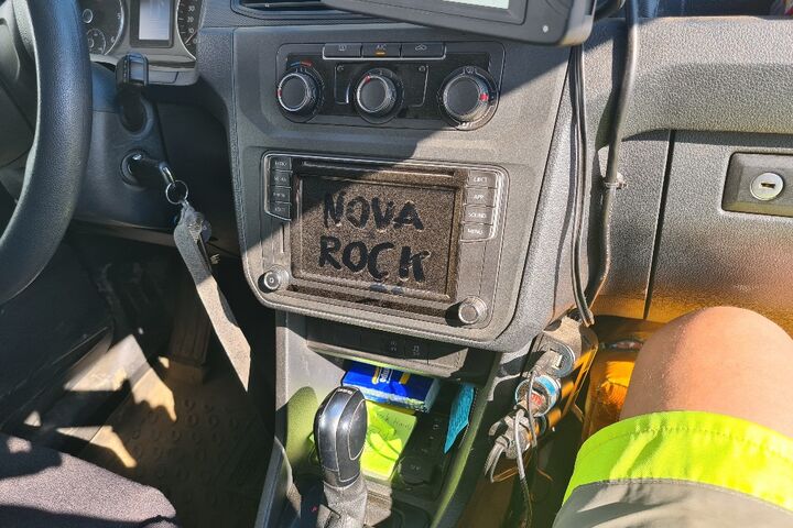 Ein Auto-Armaturenbrett mit einem in Staub geschriebenen "NOVA ROCK" auf dem Radio, daneben ein Bein in gelb-grüner Warnkleidung.