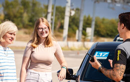Zwei Frauen stehen lächelnd neben einem silbernen Auto mit L17 Schild in der Windschutzscheibe. Vor ihnen steht ein Instruktor mit Funkgerät und erklärt etwas. Er trägt ein schwarzes T-Shirt mit gelber Fahrtechnik Aufschrift.