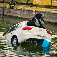 Ein Mann klettert durch die Heckscheibe eines weißen Testfahrzeuges, das halb im Wasser versunken ist