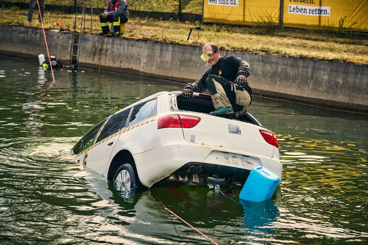 Ein Mann klettert durch die Heckscheibe eines weißen Testfahrzeuges, das halb im Wasser versunken ist