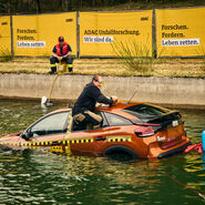 Ein Mann klettert aus dem hinteren Seitenfester eines orange-farbenen Testfahrzeuges, das halb im Wasser versunken ist