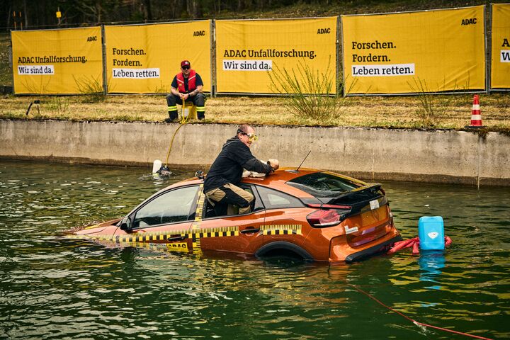 Ein Mann klettert aus dem hinteren Seitenfester eines orange-farbenen Testfahrzeuges, das halb im Wasser versunken ist