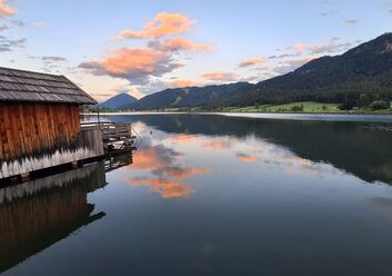 Das Foto zeigt den Weissensee, auf der linken Seite sieht man ein Holzhaus und recht einen Berg.
