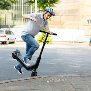 Männlicher E-Scooter-Fahrer mit Helm stürzt über Bordsteinkante
